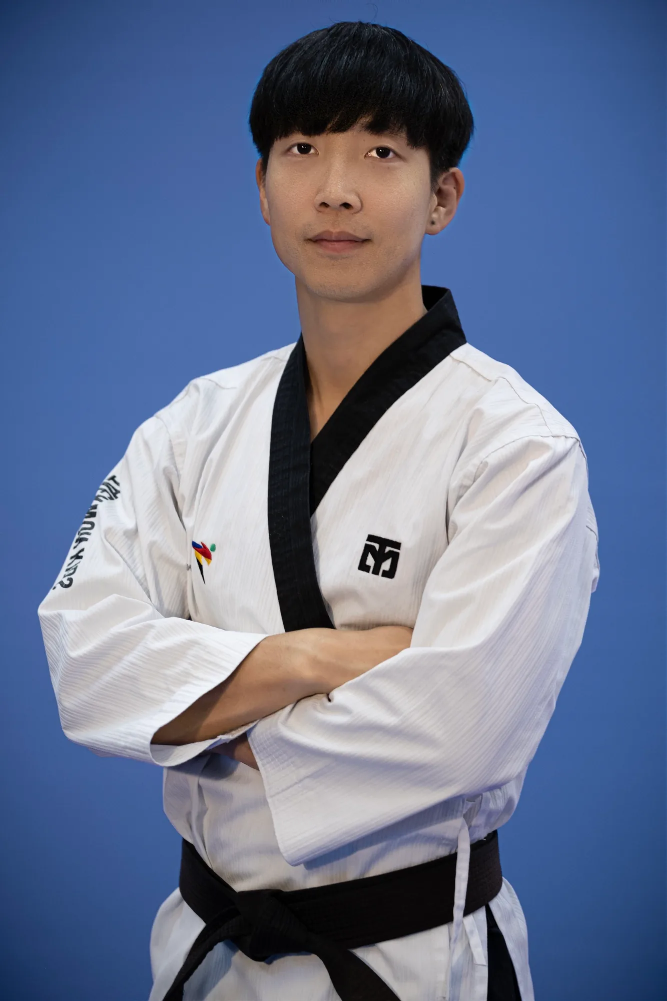 Master Joowon Lee standing in a white dobok and black belt, arms crossed, in front of a blue studio backdrop