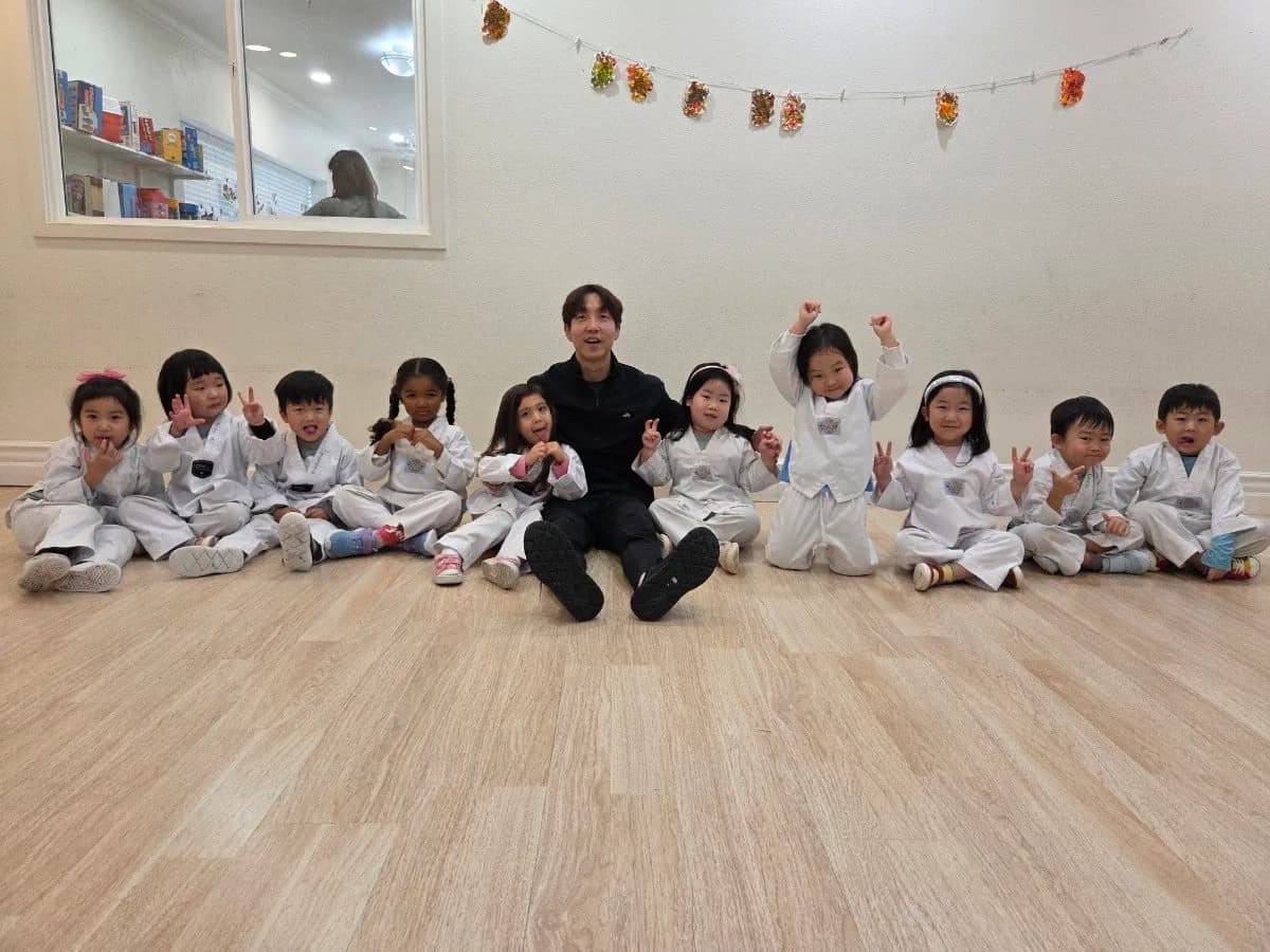 Ten young children in white doboks sitting on the floor of the training room with their instructor